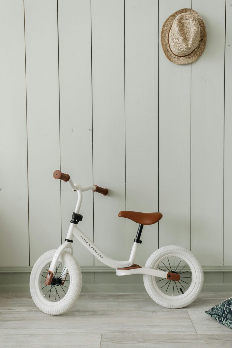 White kids' balance bike with brown accents, indoors against a stylish wall.
